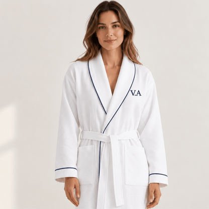 A woman wearing a white waffle-weave bathrobe with navy piping and a navy monogram 'V.A' on the chest, standing against a light beige studio background.