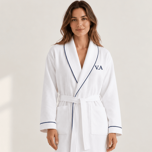 A woman wearing a white waffle-weave bathrobe with navy piping and a navy monogram 'V.A' on the chest, standing against a light beige studio background.