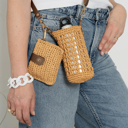 A hand-crocheted raffia bottle holder and mini clutch set in beige color with an adjustable brown strap.